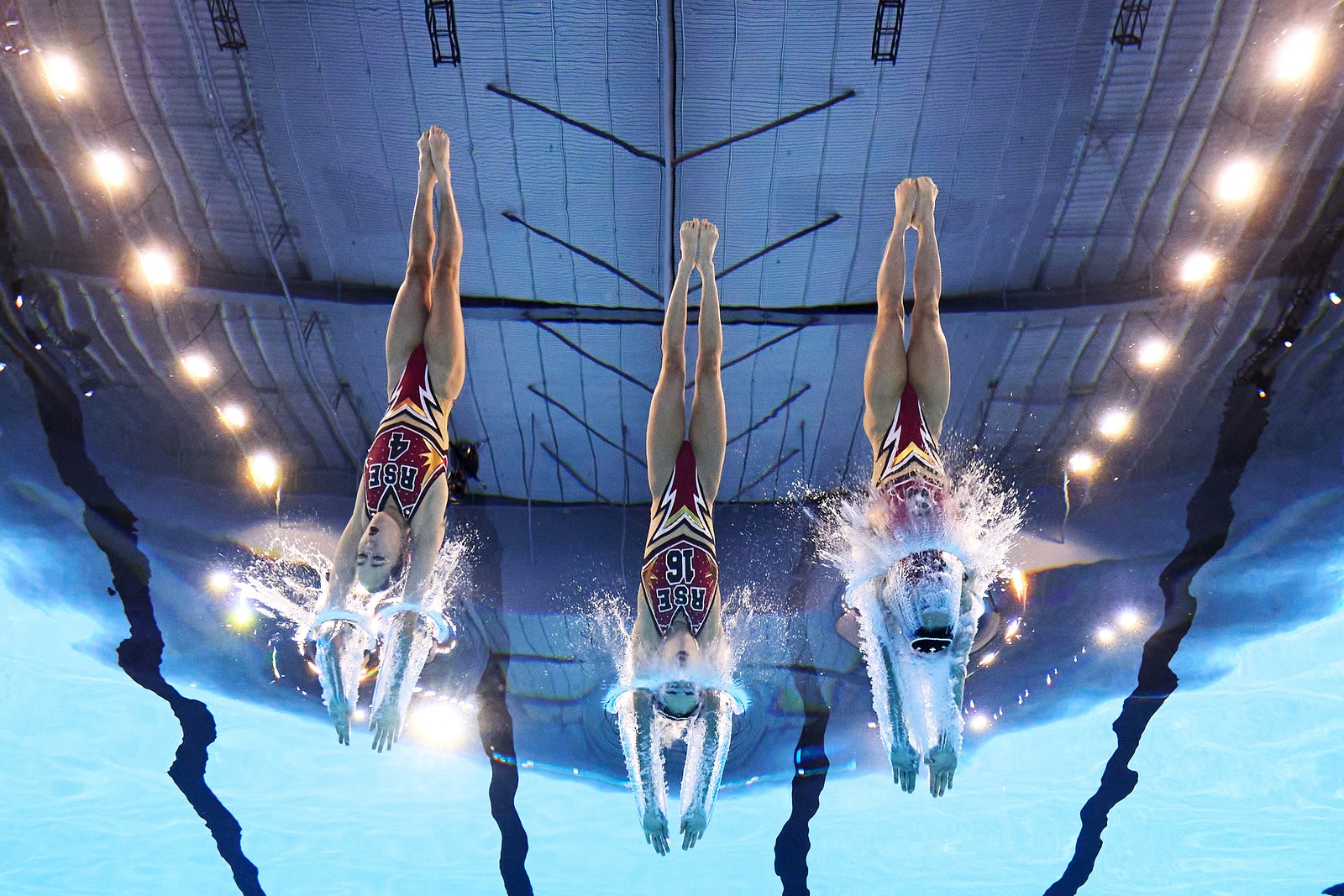 A trio of artistic swimmers dive into the pool, seen from underwater.