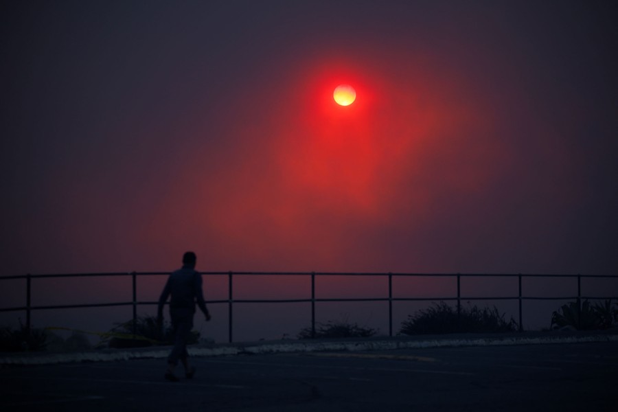 A person walks along a beach as smoke covers the setting sun.