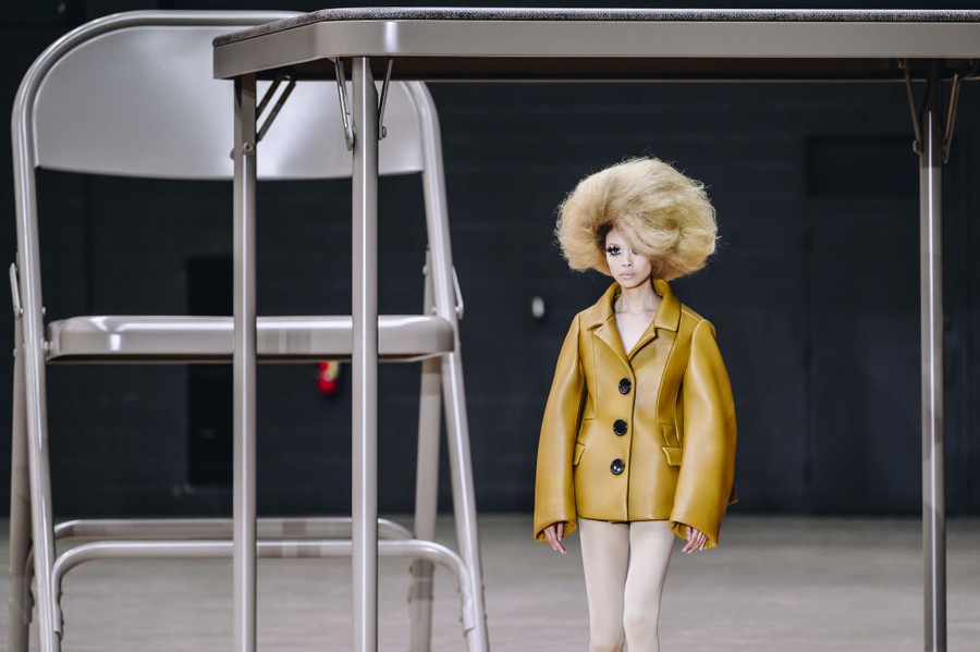 A model walks a runway in an oversize garment, passing beneath a very large folding-table-and-chair art installation.