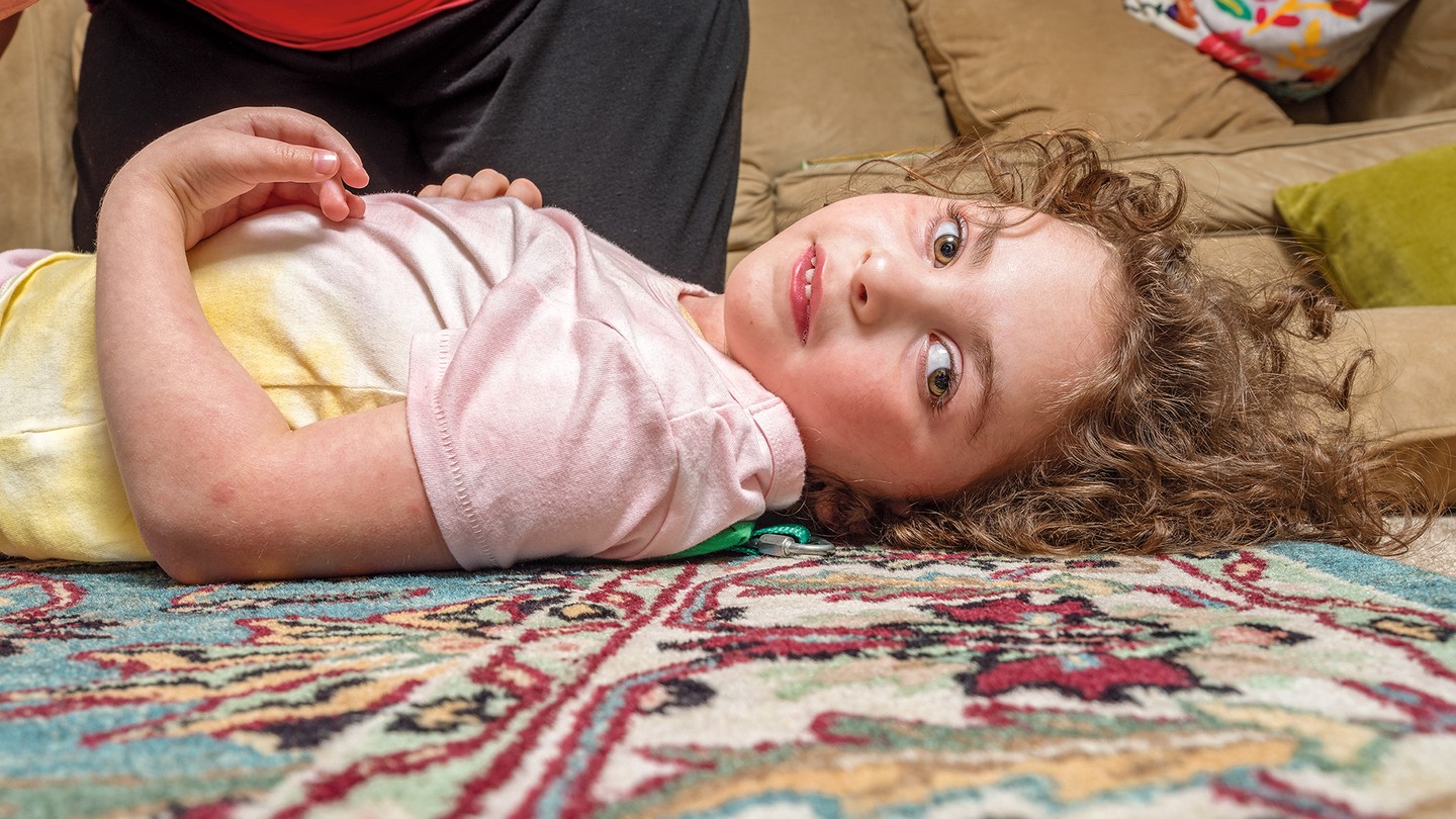 Girl with curly hair lying on colorful carpet with head turned toward the camera, with someone kneeling next to her and couch in background