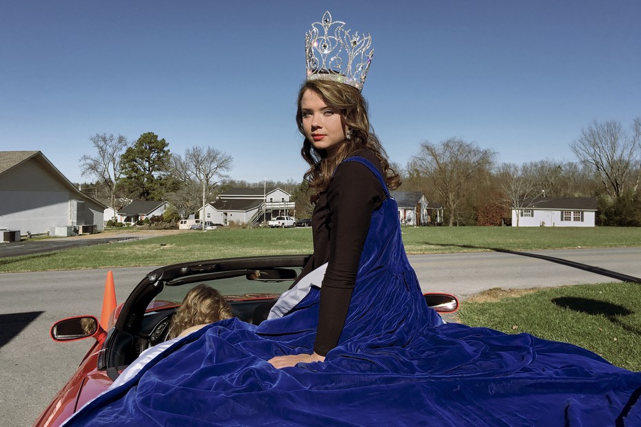 A young woman turns to the camera sitting on the back of a convertible car wearing a gown and tiara.