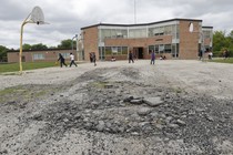 Kids play on a broken basketball court. The net on the hoop is missing. A brick school is in the distance. 