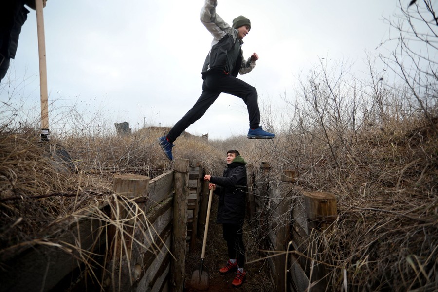 One teen stands in a trench with a shovel while another leaps across the trench above.