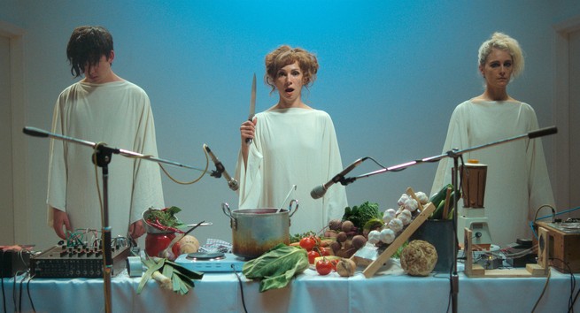 Three people dressed in white standing in front of a table filled with food and recording equipment
