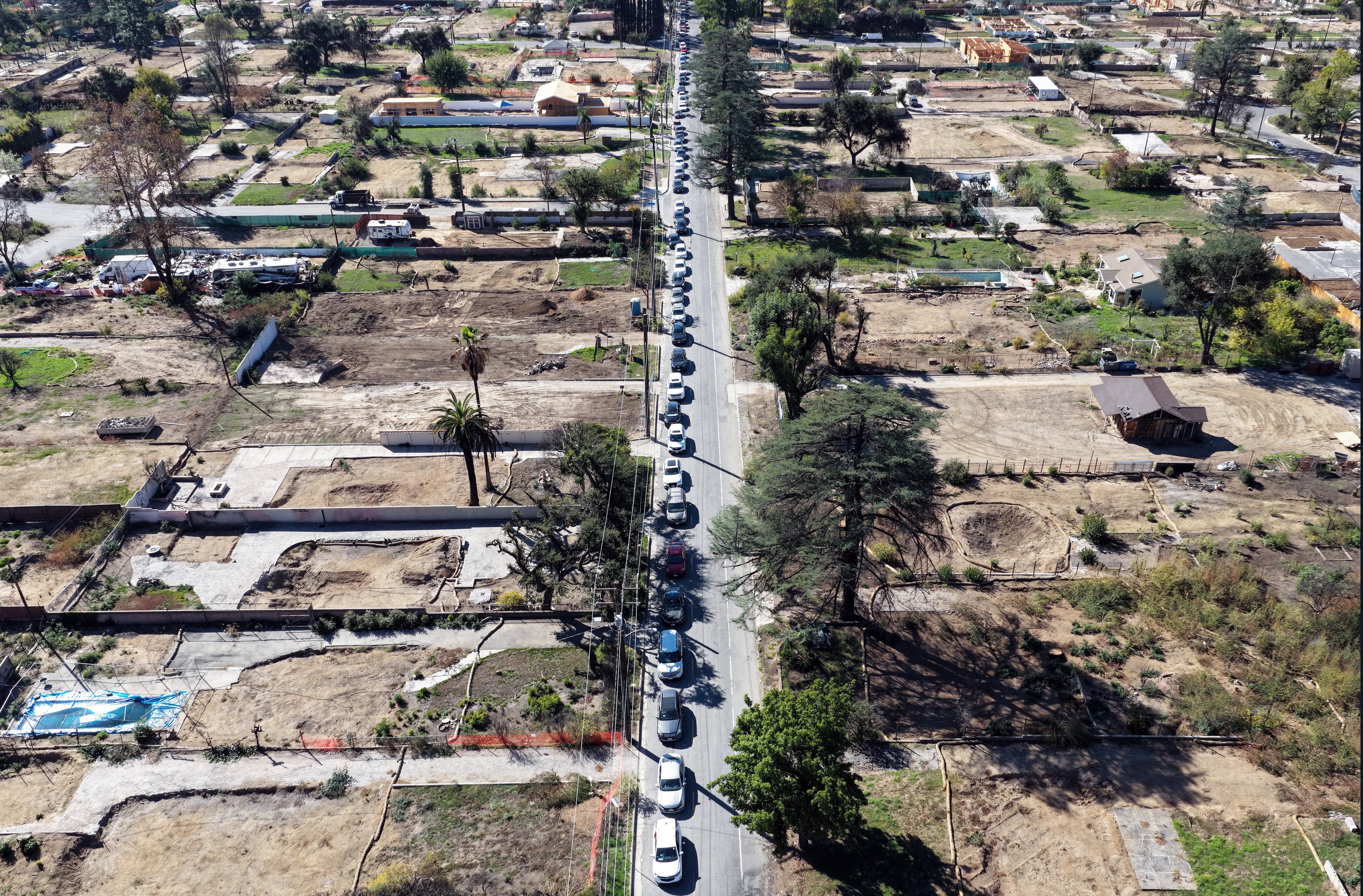 An aerial view of a long line of cars on a street that passes through many empty lots in a residential area affected by a wildfire months ago.