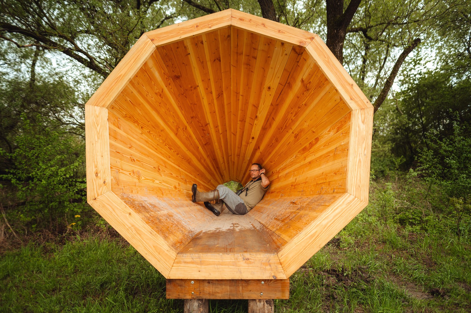 A person sits inside a giant wooden cone, with their hand to their ear, listening.