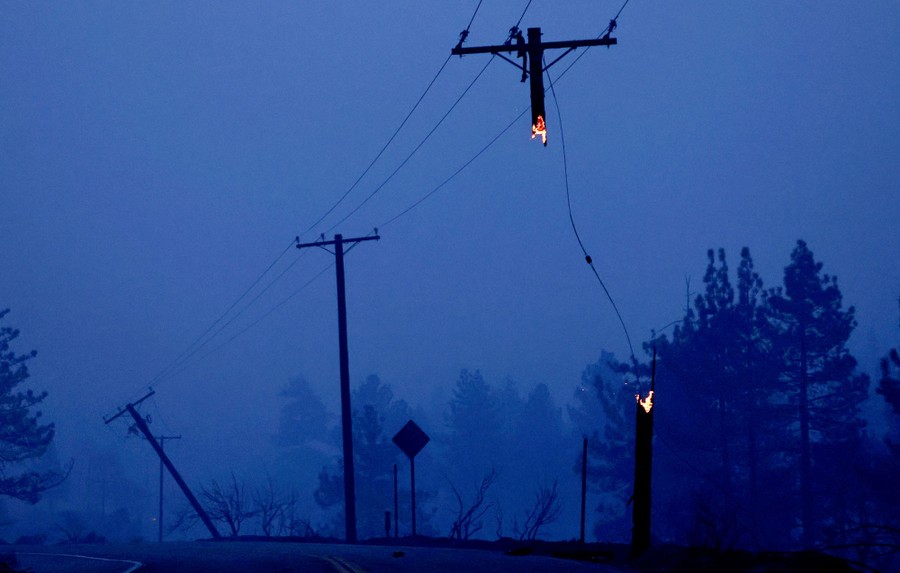 A fire-damaged set of power poles, including one pole that has burned through, with the middle section missing. The top, still burning, hangs from wires.