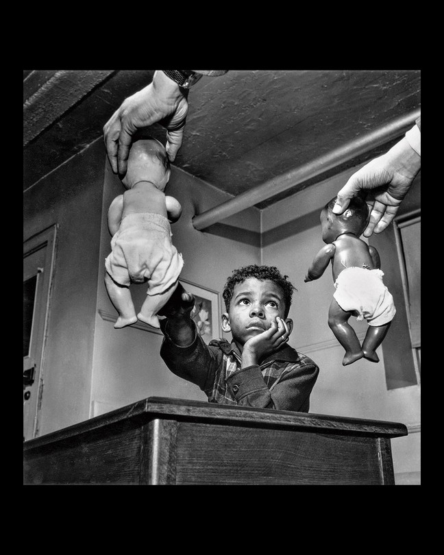 Black and white photo of two hands offering dolls to child seated at desk, child reaching for the white doll while looking at the Black doll