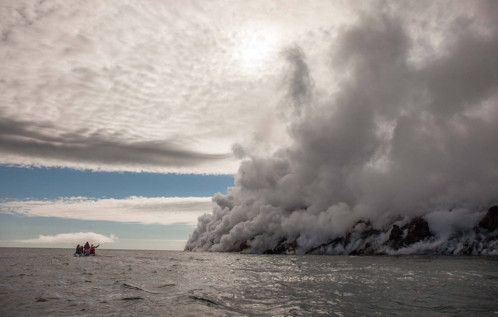 Visitors in a small boat observe a volcanic flow reaching the ocean, sending plumes of steam into the air.