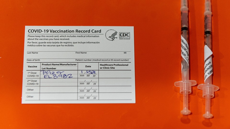 A photo of a COVID-19-vaccination card next to two syringes filled with Pfzier-BioNTech vaccine doses