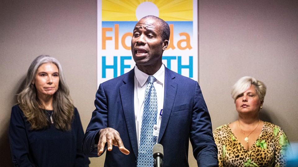 Florida Surgeon General Joseph Ladapo delivers a speech in a blue suit as two women watch behind him.