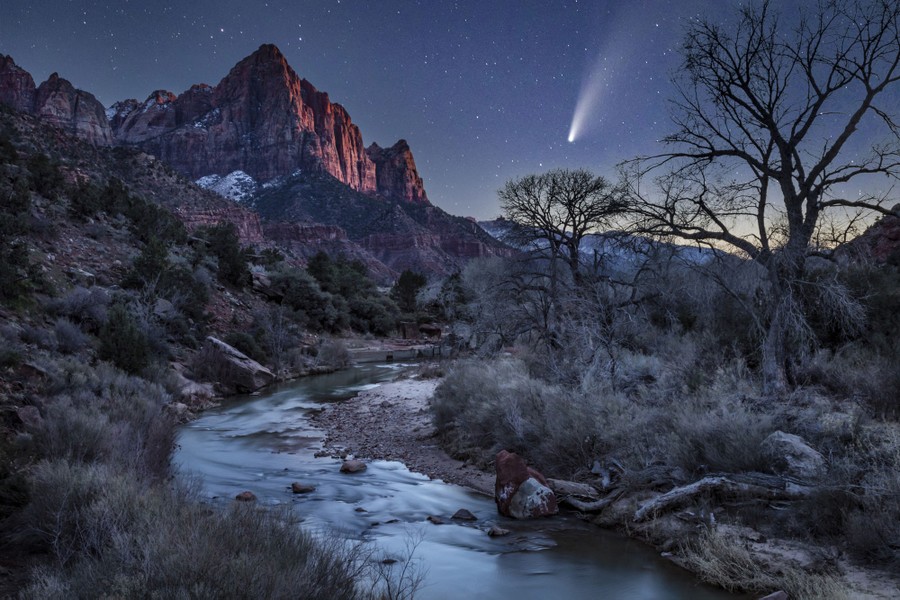 A comet appears in the night sky above a desert valley with a small river flowing through it.