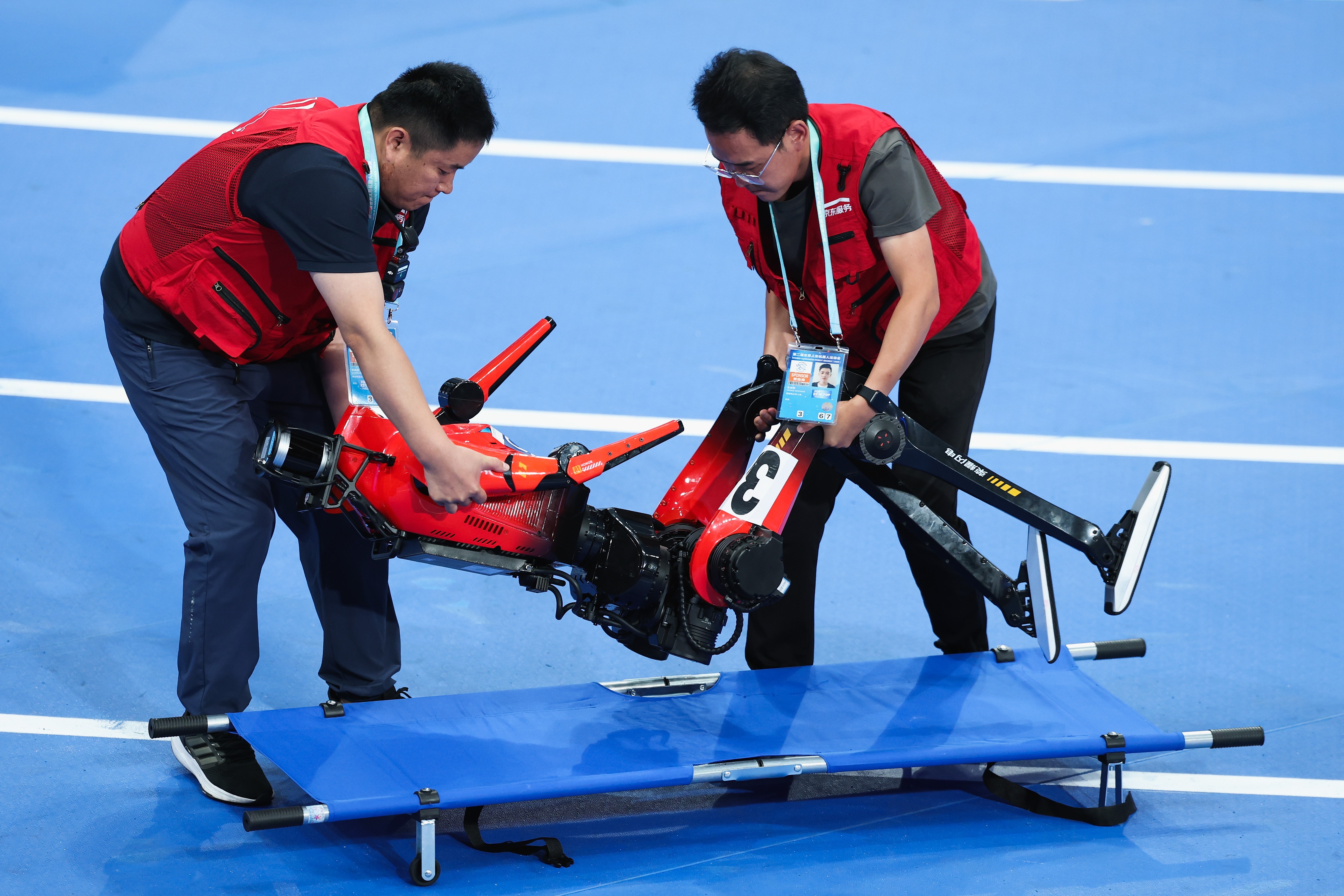 Two people lift a fallen humanoid robot onto a stretcher.