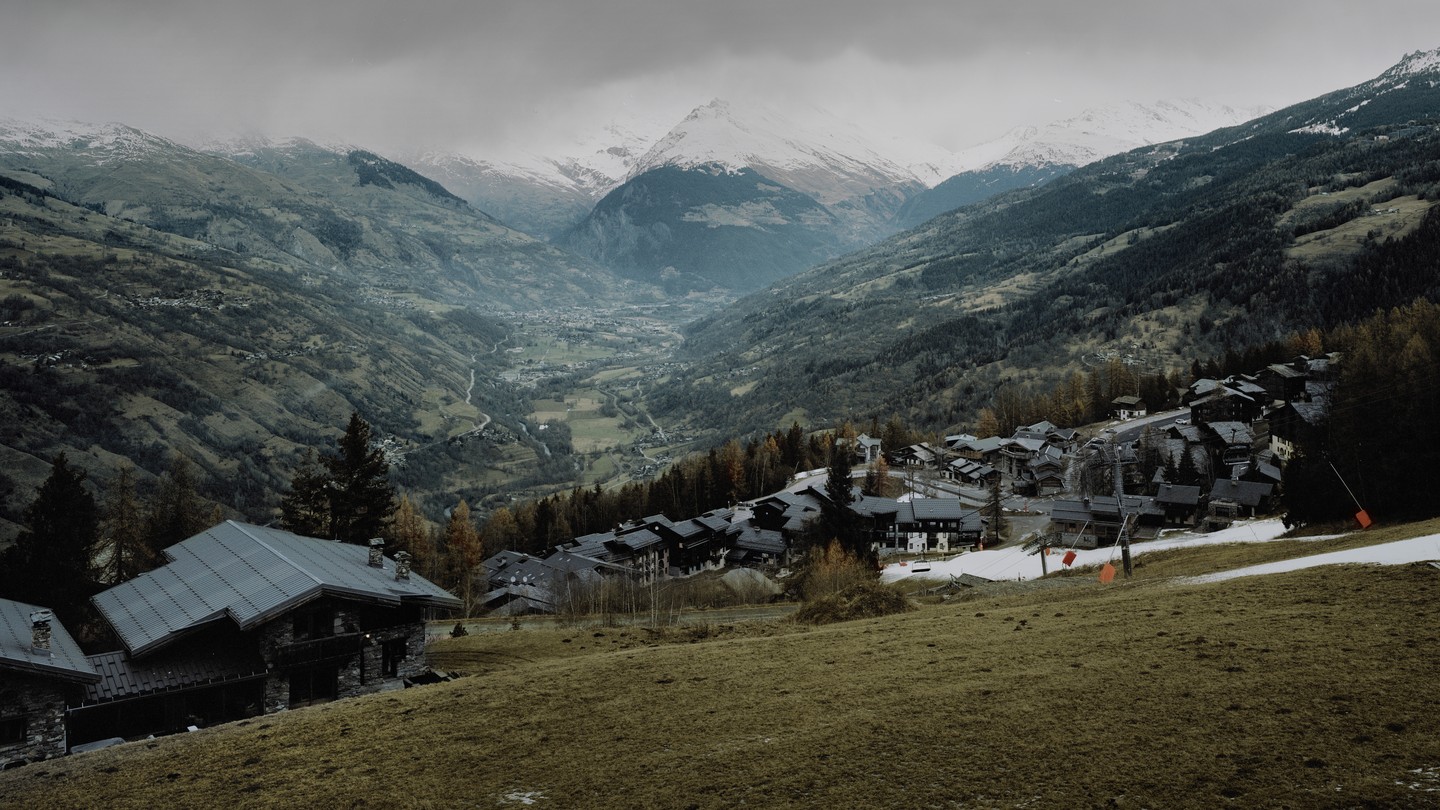 Photo of a mountainside with buildings, some snow, clouds overhead, and a valley below