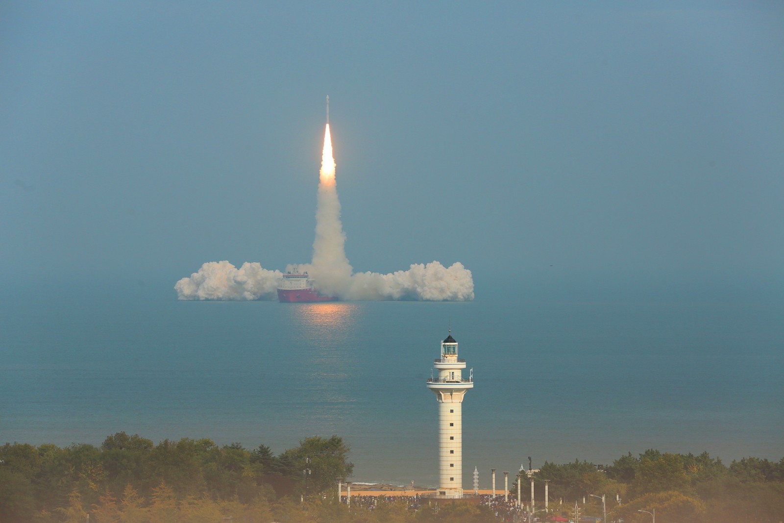 A rocket lifts off from the deck of a ship, seen from a distance with a lighthouse in the foreground.