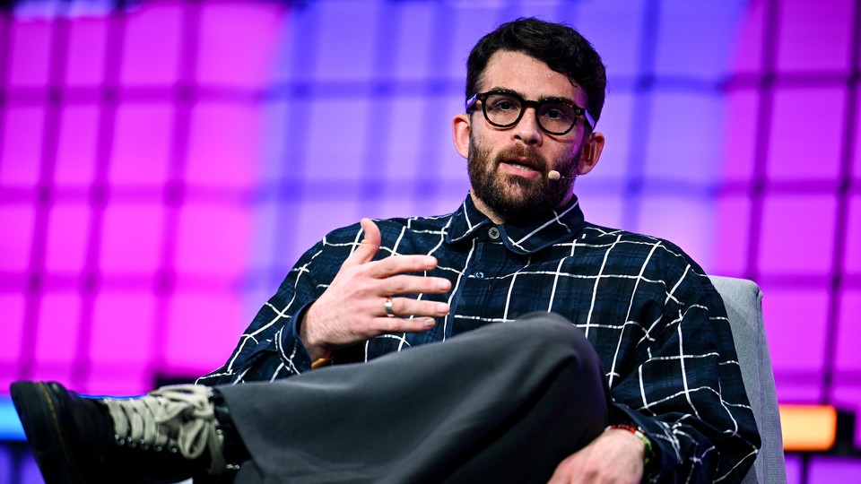 Photograph of Hasan Piker speaking with a microphone attached to his face. He is seated in a gray chair with his legs crossed.