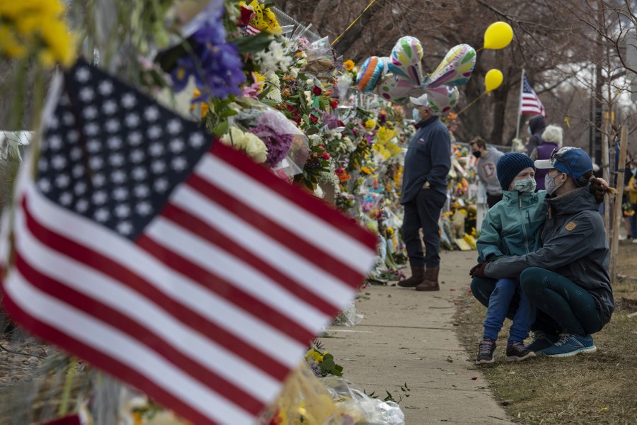 People visit a long wall covered in flower bouquets, balloons, and flags.