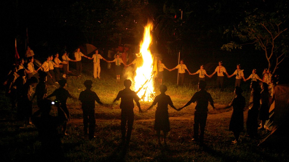 Children in scout uniforms hold hands around a bonfire