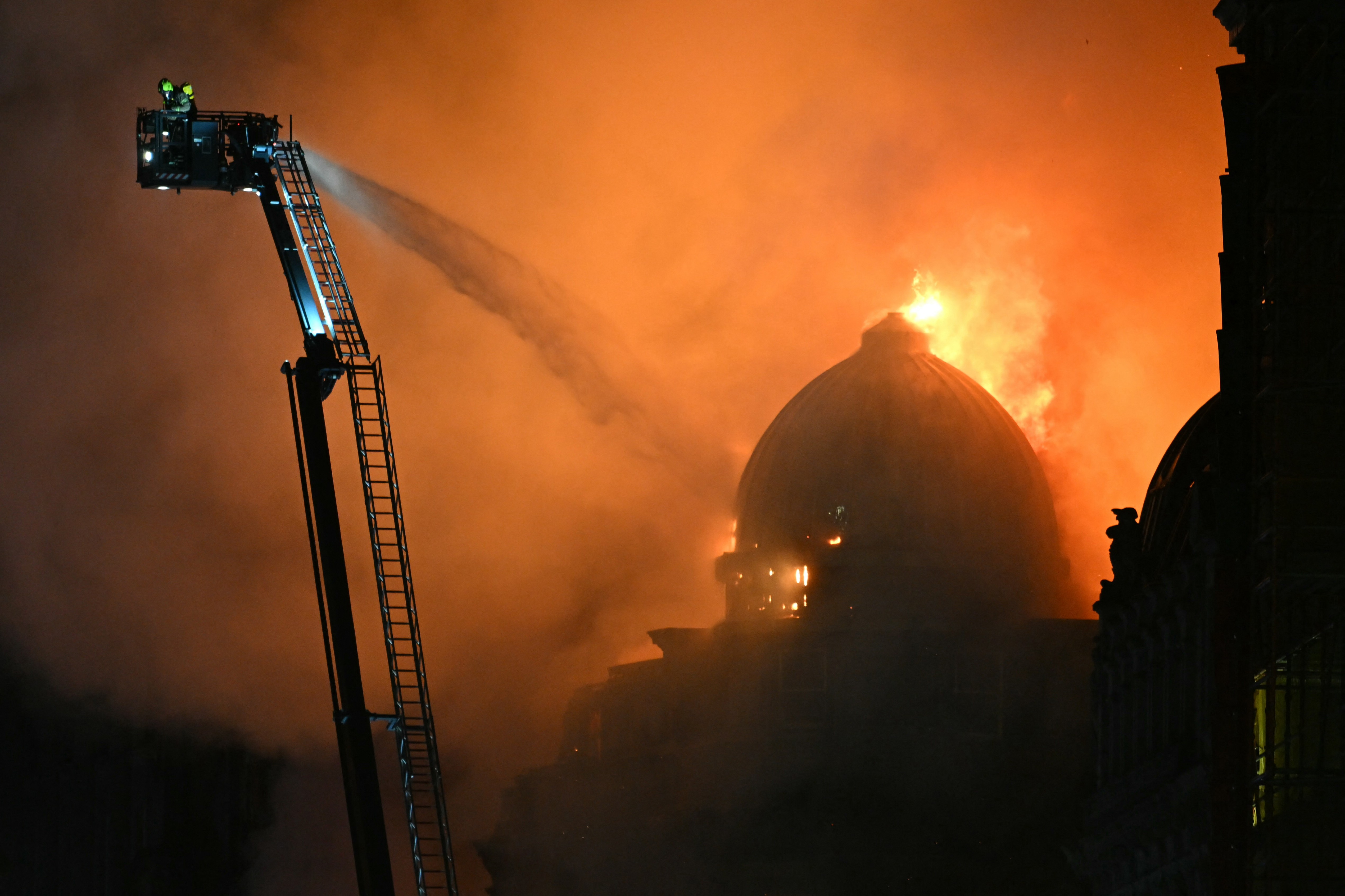 Firefighters on a tall extended ladder rig spray water on a fire burning in a large domed building at night.