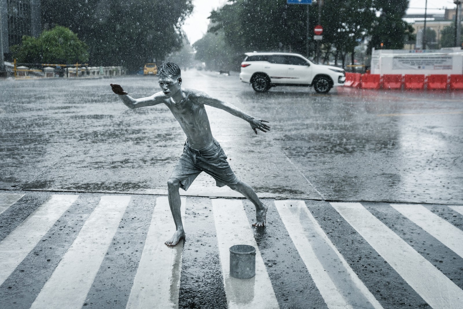 A silver-painted street performer poses on a crosswalk during a rain storm.