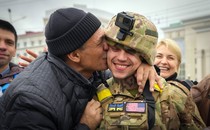 A civilian kisses a smiling soldier as others look on.
