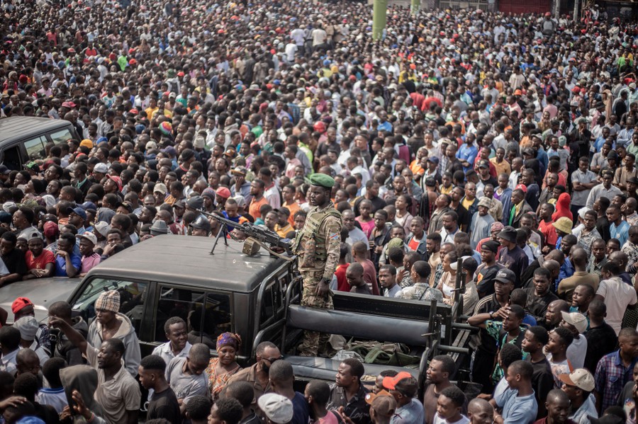 A large crowd of civilians stand alongside a a pickup truck with an armed soldier standing in its bed.