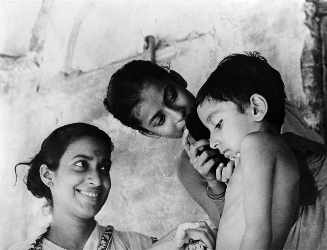 A black and white photo of young boy and his older sisters who are brushing his hair and smiling at him
