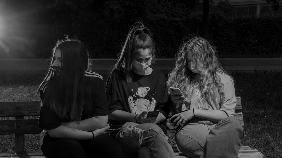 Three teenagers wearing masks look at their phones in a park