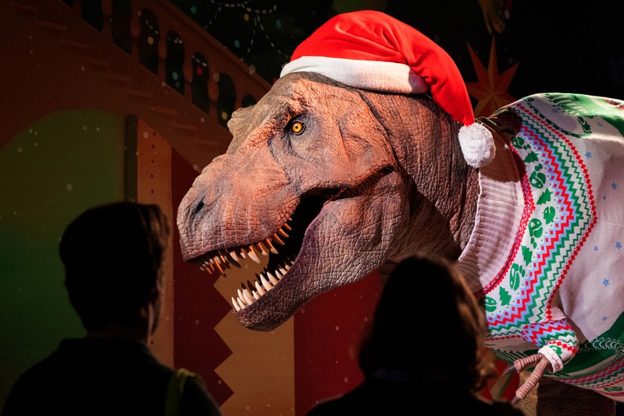 People look at a life-size T. rex sculpture dressed in a Santa hat and festive sweater.