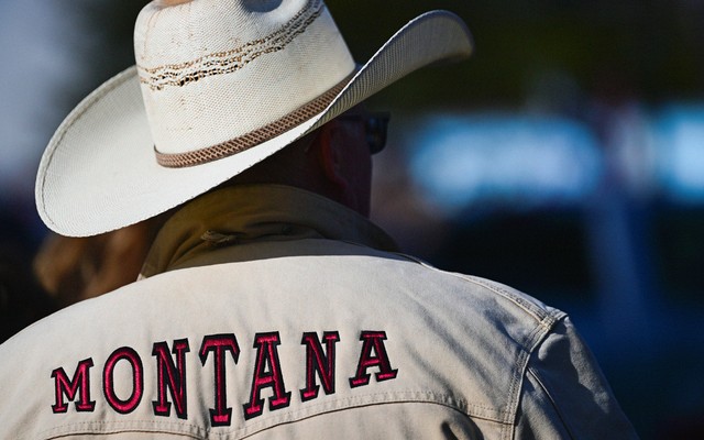 A photograph of a man from behind, wearing a white cowboy hat and a white jacket that says MONTANA in red font across the shoulders