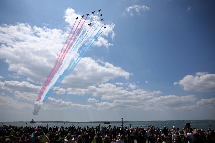 A formation of 10 fighter jets flies above a crowd beside the ocean, leaving red, white, and blue smoke trails.