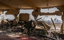 A soldier walks through a blown out interior in Gaza, ceiling fans and carpets visible under rubble but walls blasted through.