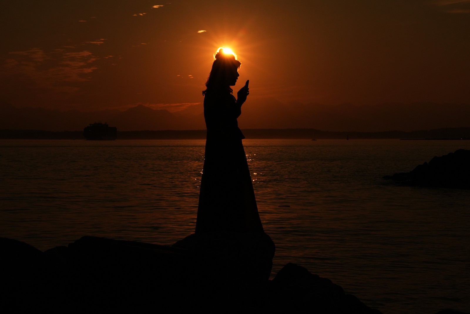 A woman is silhouetted against the setting sun while holding up her phone.