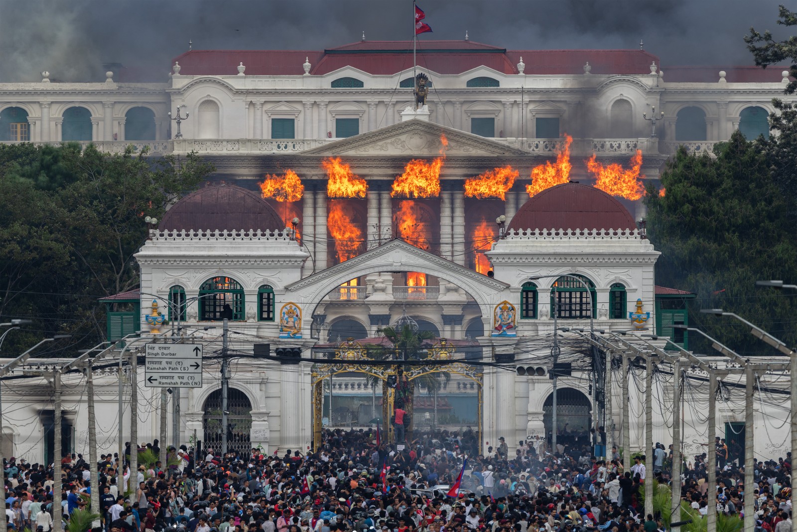 Fire and smoke rise from a large government building as protesters crowd beneath a gate.