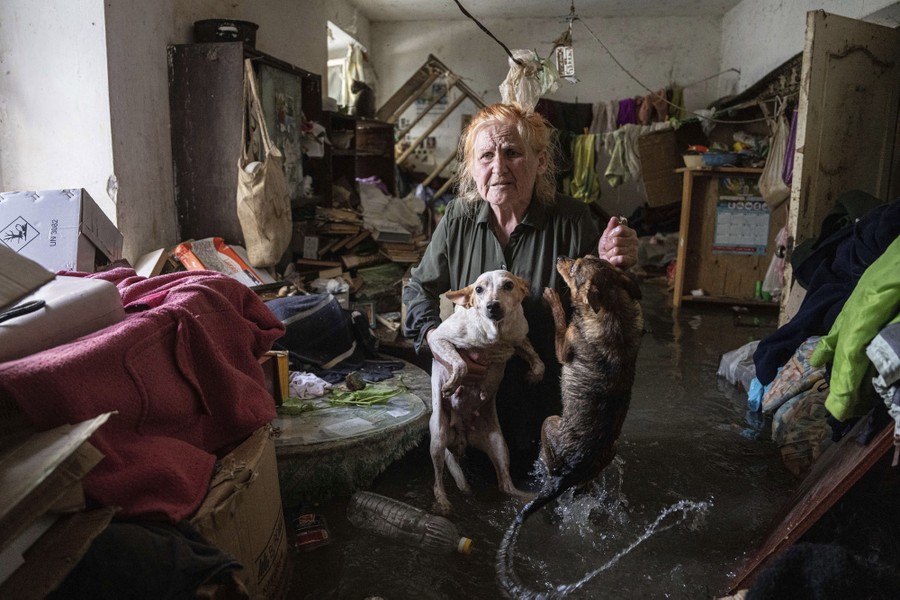 A woman stands inside a room in knee-deep floodwater, holding two small dogs.