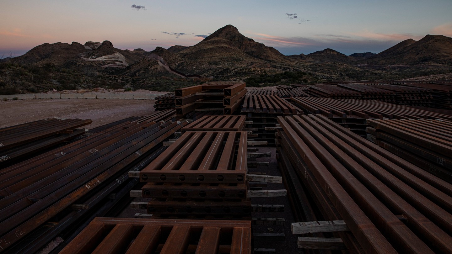 Stacks of steel bollards sit in a desert. In the background, a blasted mountain and Donald Trump's unfinished wall.