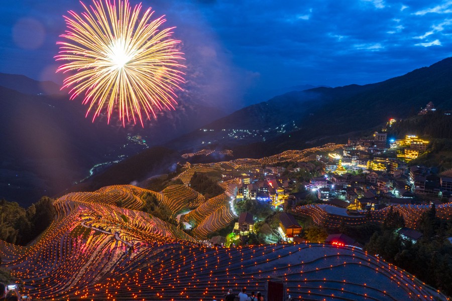 Fireworks explode above terraced fields on a steep hillside decorated with lights.