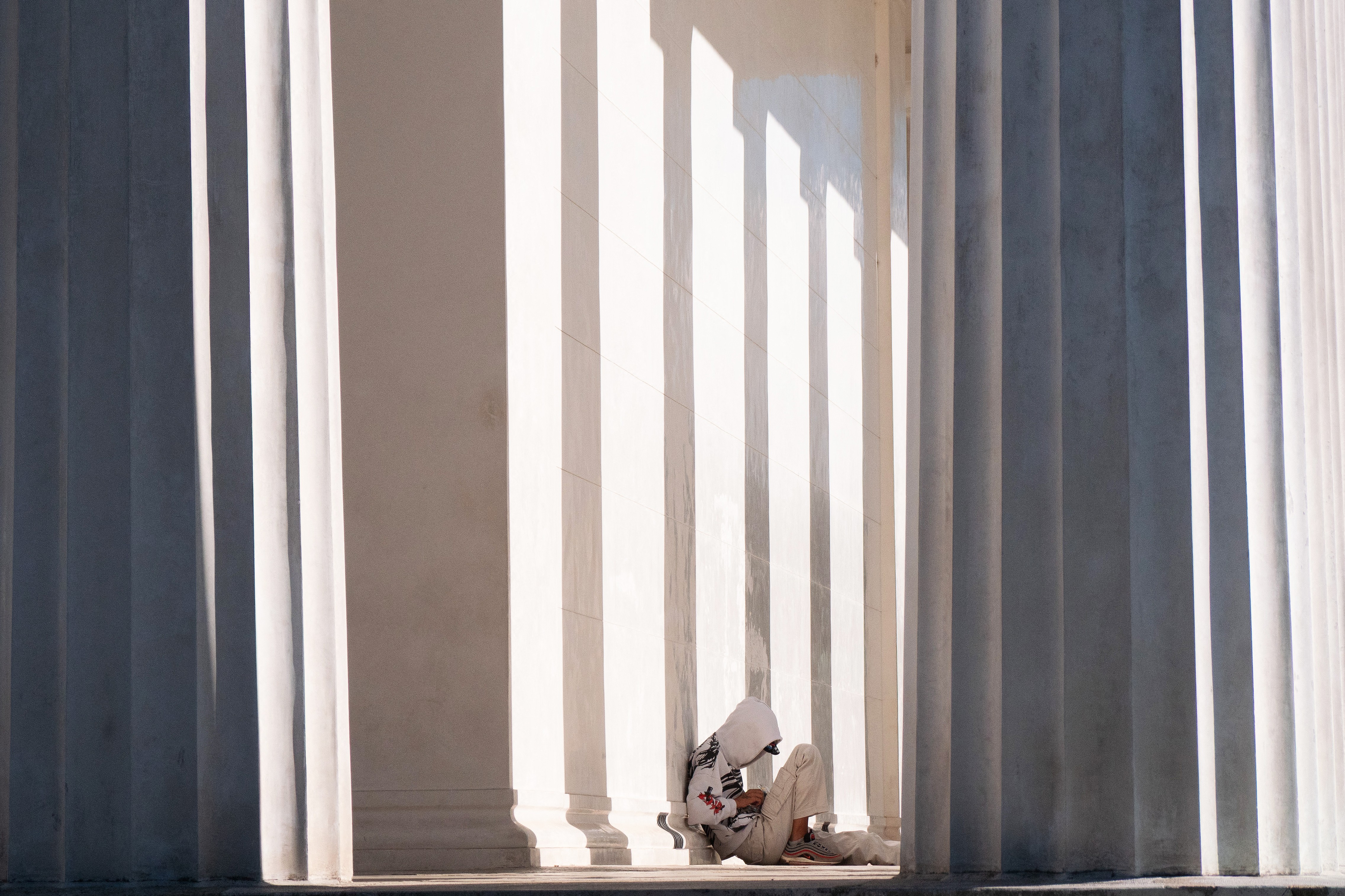 A person wearing a hoodie sits on the floor beside a building with many tall columns.