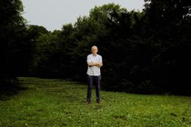 A color photograph of a white man standing with arms crossed on a green lawn surrounded by dark trees