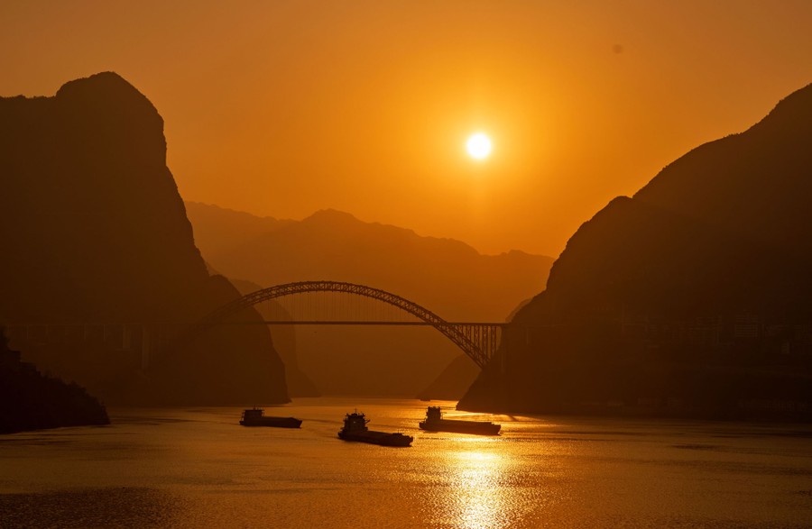A low sun is seen over a broad river gorge, a road bridge, and several barges.