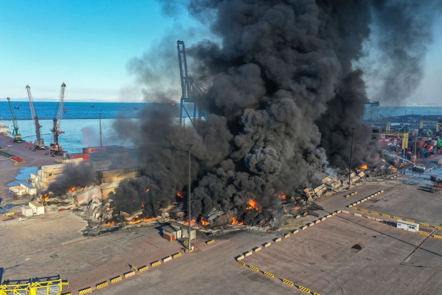 Black smoke rises from a large fire burning in a port facility.