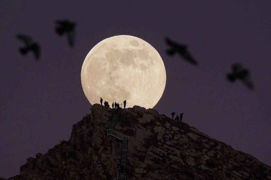 A small group of people in silhouette atop a rocky hilltop, in front of a full moon