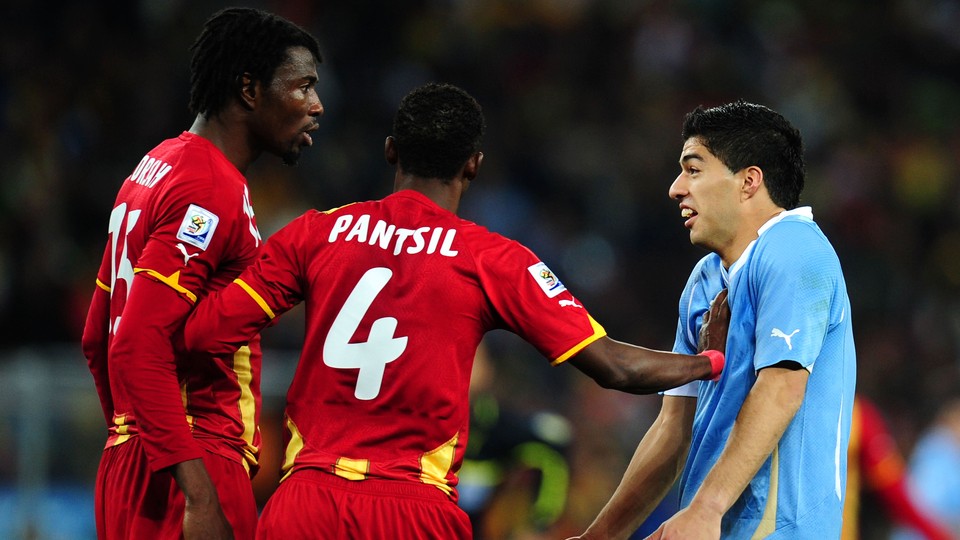 A soccer player in red stands between a teammate and another player in blue.