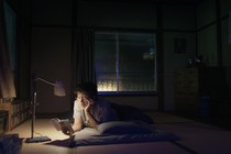A photograph of a person laying on the floor of a cozy bedroom at night with a lamp illuminating the book the person is reading