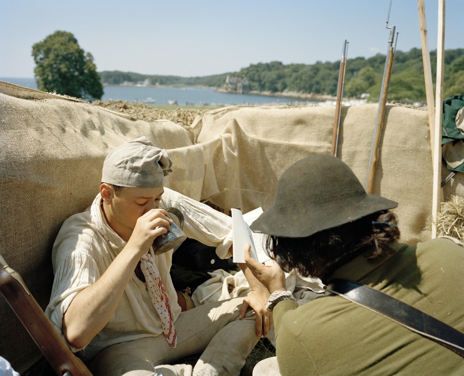 photo of two reenactors dressed as American soldiers resting, one taking a drink of water and the other reading, during a pause in the reenactment, with the shore in the background
