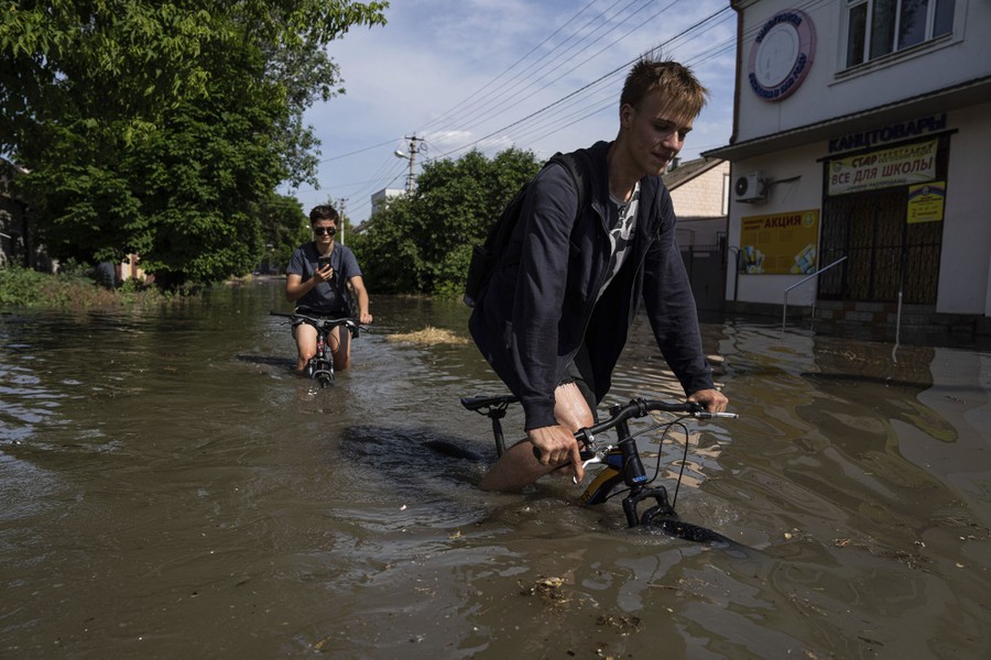 Two people try to ride their bikes through deep floodwater.
