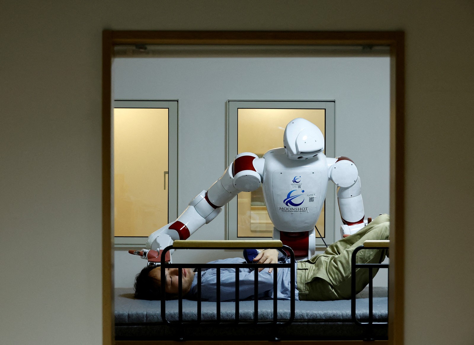A humanoid robot prepares to roll a human patient onto their side on a bed.