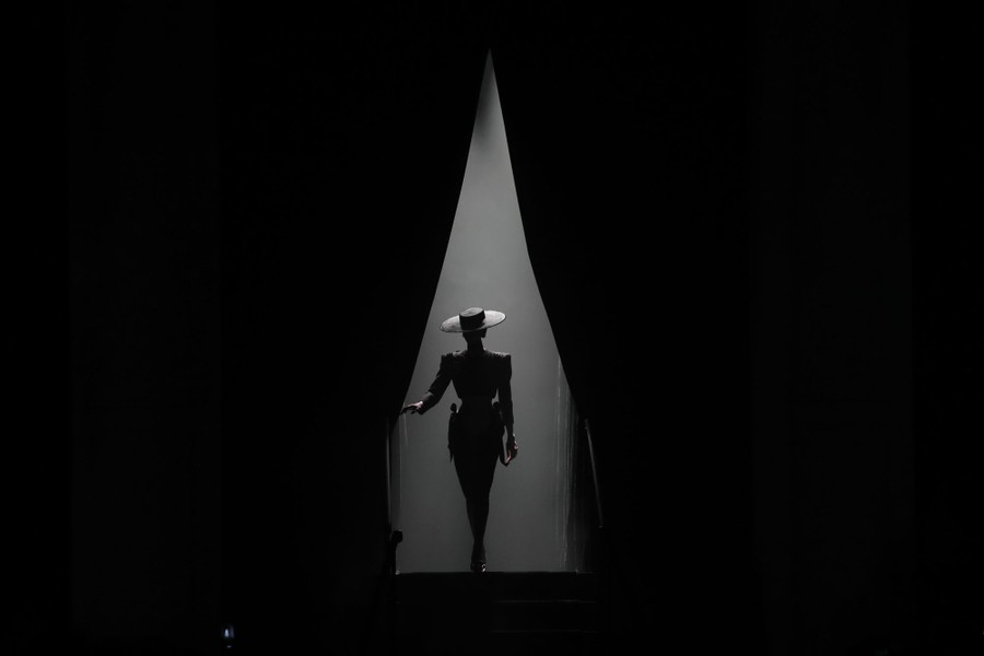 A model wearing a broad-brimmed hat walks through a draped opening.