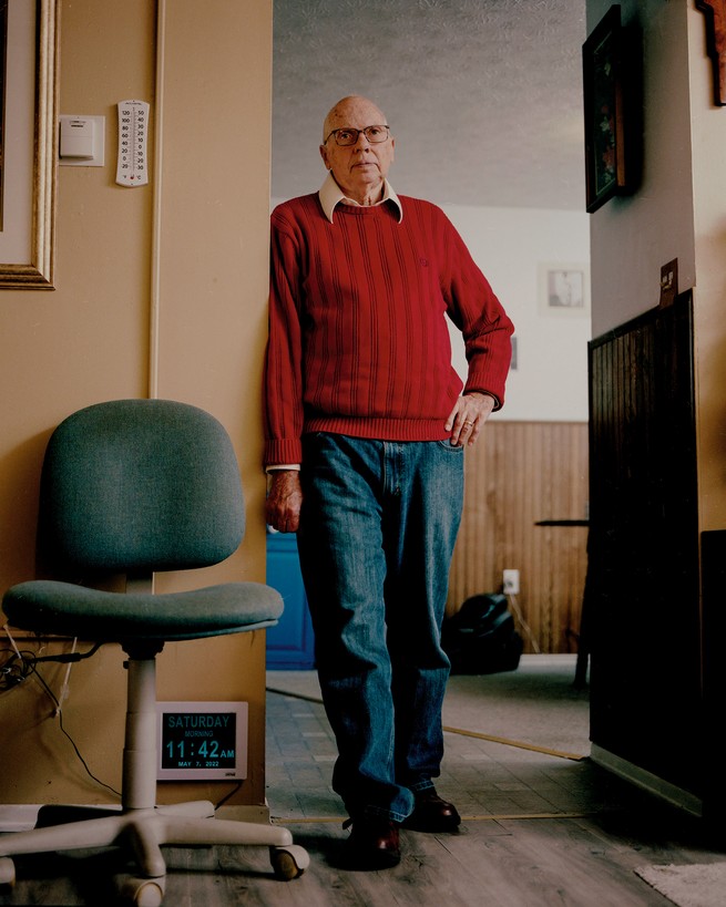 photo of man wearing collared shirt under red sweater and jeans standing with his hand on his hip in a doorway