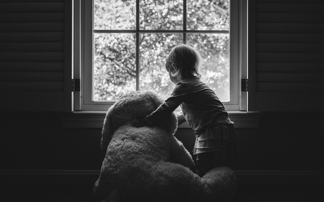 A young girl holding a large teddy bear stares out the window.
